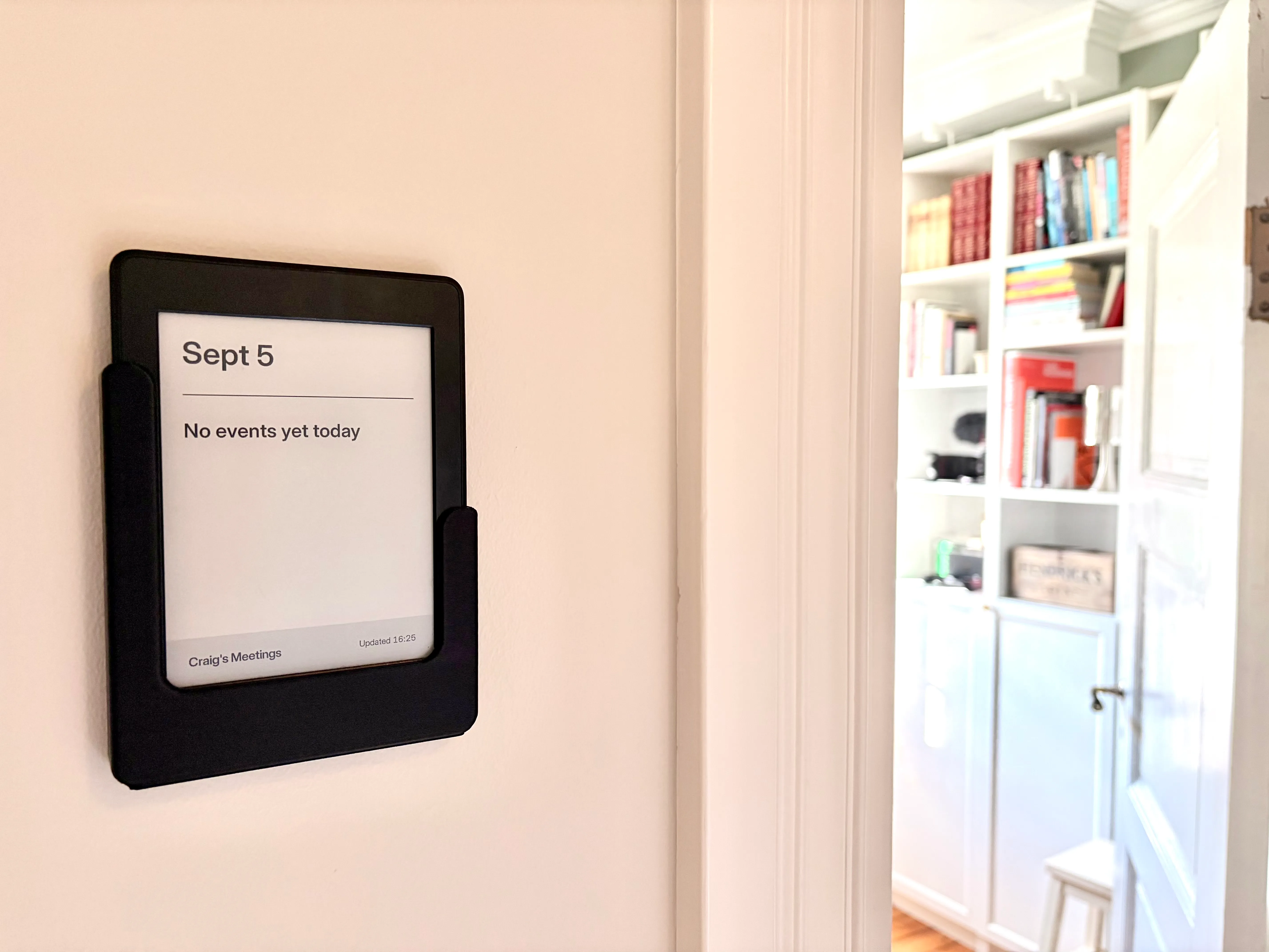 A Kindle in a black wall mount beside an open office door. The e-ink screen reads Sept 5 above a divider, then No events yet today, with a grey footer bar reading Craig's Meetings and Updated 16:25. Bookshelves are visible through the door.