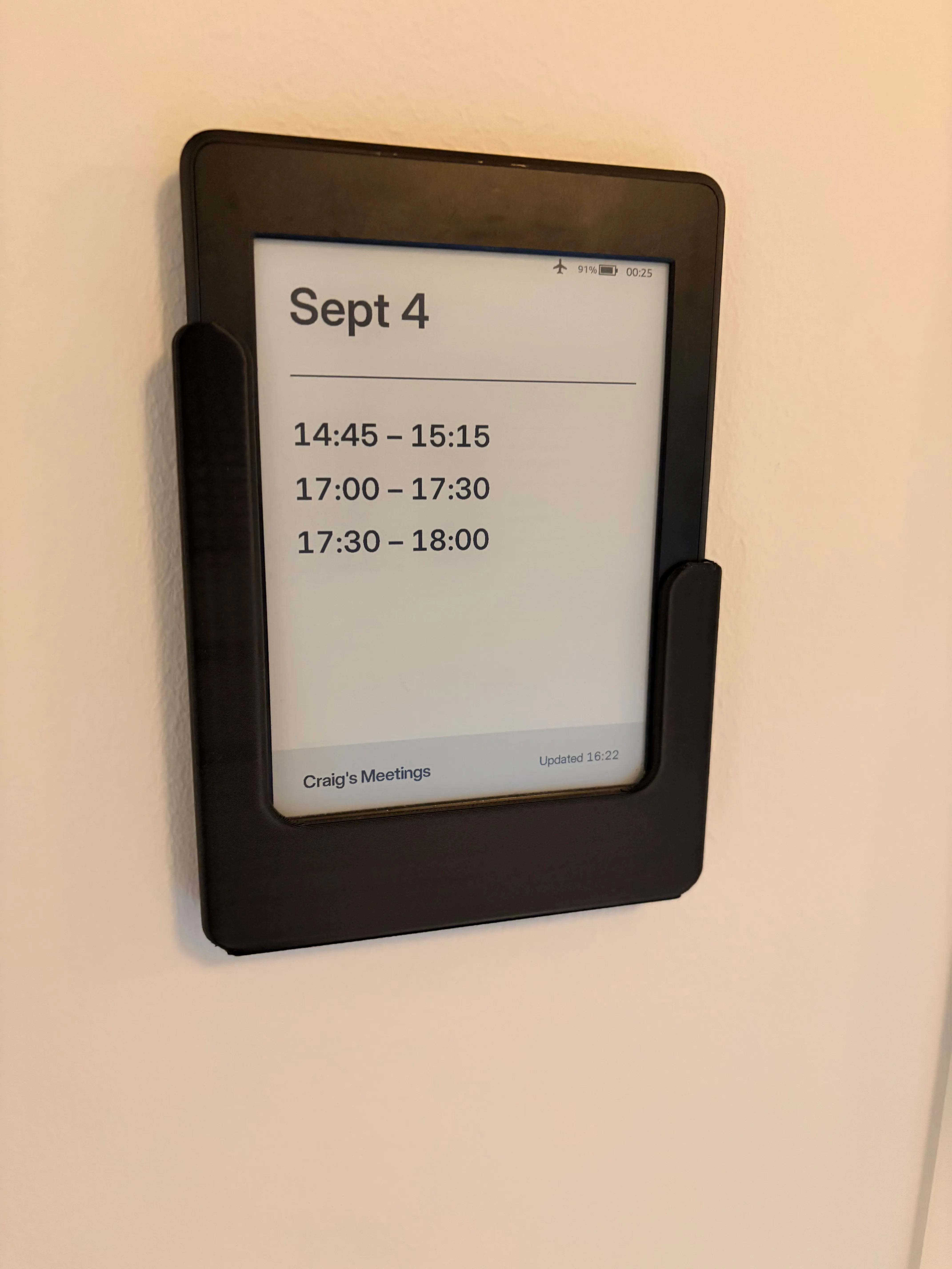 A Kindle Paperwhite in a black 3D-printed wall mount on a plain wall. The screen reads Sept 4 above a divider, then three meeting times in large type: 14:45 to 15:15, 17:00 to 17:30 and 17:30 to 18:00. A grey footer bar reads Craig's Meetings and Updated 16:22.