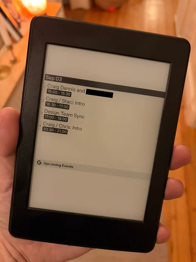 A Kindle held in one hand, showing TRMNL's stock Google Calendar plugin rotated upright. Sep 03 in a black header bar, then four numbered meetings with titles and times in small type, one attendee name blacked out, and a Google logo with Upcoming Events at the bottom.